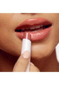 Rossetto in una delicate tonalità nude applicato con le dita, con un elegante tubo bianco. Le labbra hanno una finitura lucida e un colore naturale.