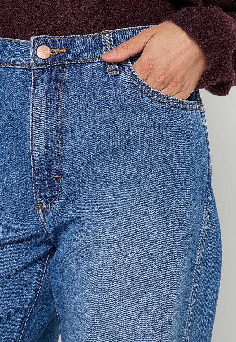 Jasnoniebieskie dżinsy o wysokim stanie z przednią kieszenią i kontrastowym pomarańczowym przeszyciem. Widoczny detal z miedzi.