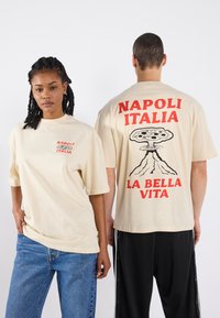 Beige bomulls t-skjorte med rød tekst "NAPOLI ITALIA" på forsiden og et grafisk motiv av en vulkan med pizza på baksiden. Løs passform.