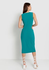 Tschickribbed Midi-Kleid mit Cut-out-Seitendetails, figurbetontem Schnitt, rundem Halsausschnitt und subtiler Textur. Kombiniert mit weißen Riemchenschuhen.