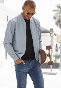 Light blue bomber jacket with a zippered front, ribbed cuffs, worn over a black t-shirt, paired with blue jeans and holding a skateboard.