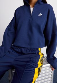 Sudadera recortada de color azul marino con cuello alto y media cremallera, que presenta un logo de Adidas bordado en blanco. Combinada con pantalones de chándal en azul marino y amarillo.