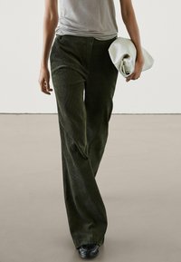 Pantalon large en velours côtelé vert foncé avec une finition texturée, porté avec une chemise grise et tenant une pochette blanche. Design simple sans motifs visibles.