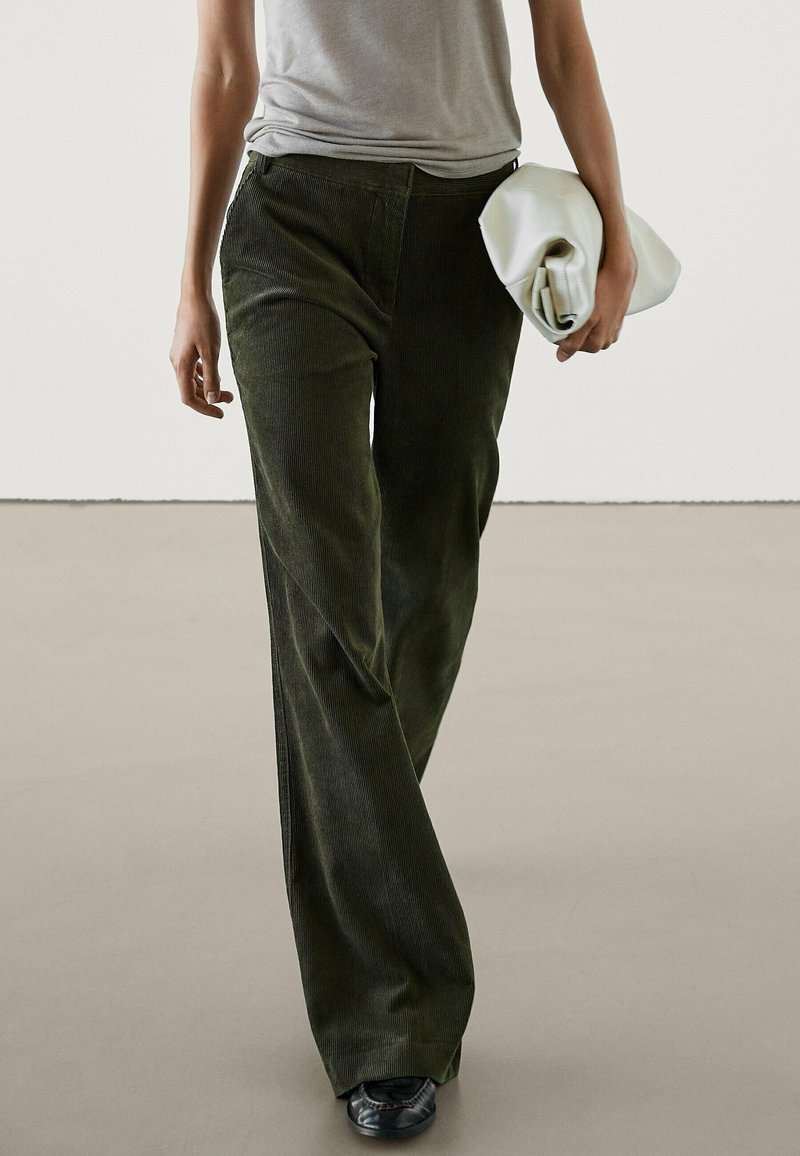 Pantalon large en velours côtelé vert foncé avec une finition texturée, porté avec une chemise grise et tenant une pochette blanche. Design simple sans motifs visibles.