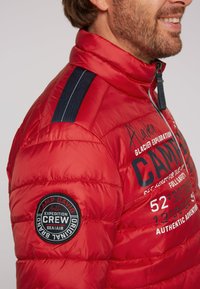 Giacca a vento rossa con colletto alto, righe blu navy sulle spalle e un patch circolare sul maniche con dettagli testuali.