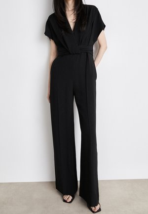 Kobieta ubrana w czarny, krótki kombinezon z zakładanym dekoltem, szerokimi nogawkami i czarnymi sandałami z odkrytymi palcami, stoi na tle białej ściany.