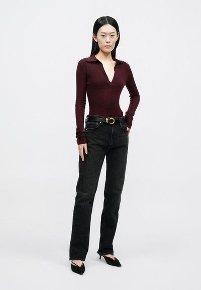 Γυναίκα όρθια με τα χέρια στις τσέπες, φοράει μπορντό μακρυμάνικη μπλούζα, μαύρα τζιν με ζώνη και μαύρες γόβες μυτερές, σε λευκό φόντο.