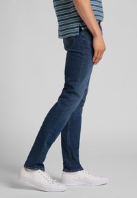 Mörkblå denimjeans med slim fit, med en klassisk femfickdesign, bärs med vita canvas-sneakers.