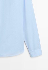 Jasnoniebieska koszula z długim rękawem, wykonana z teksturowanej tkaniny. Posiada zaokrąglony dół oraz zapięcie na guzik przy mankietach, co umożliwia regulację dopasowania. Minimalistyczny design.