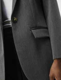 Blazer gris cintré avec une fermeture à un bouton, un col à revers et une poche passepoilée sur le côté gauche.