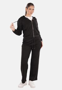 Felpa nera con zip, cordoncino e due tasche, abbinata a pantaloni neri coordinati, realizzati in tessuto morbido, indossati con sneakers bianche.