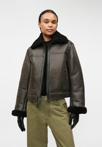 Barna bőr, rövid fazonú dzseki fekete prém gallérral és mandzsettákkal, elől cipzárral és két oldalsó zsebbel. Simára kidolgozott felület, kontrasztos varrásokkal.