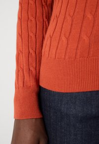Cable-knit sweater in burnt orange featuring ribbed cuffs and hem, paired with dark denim jeans, showcasing texture and color contrast.