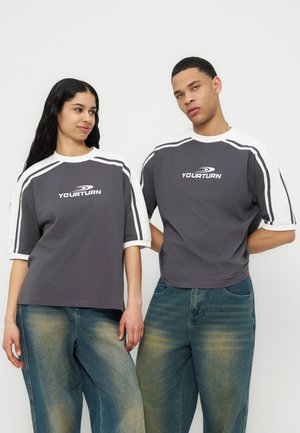 Δύο μοντέλα φορούν ταιριαστά γκρι και λευκά μπλουζάκια με το λογότυπο "YOURTURN" και ξεθωριασμένα μπλε τζιν παντελόνια, στέκονται μπροστά σε ένα λευκό φόντο.