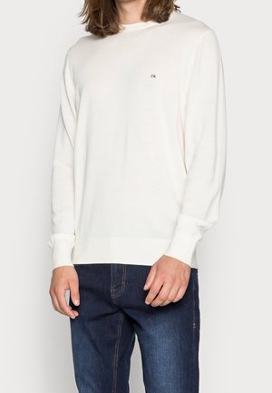 Cremfarbener Strickpullover mit langen Ärmeln, Rundhalsausschnitt und kleinem gesticktem Logo in der Mitte. Kombiniert mit dunkelblauen Jeans.