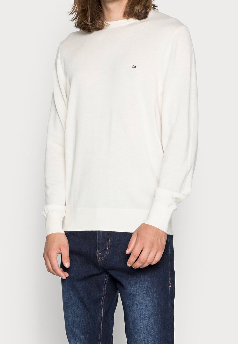 Maglione lavorato a maglia color crema con maniche lunghe, colletto a girocollo e un piccolo logo ricamato al centro. Abbinato a jeans blu scuro.