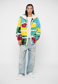 Fuzzy multi-colored jacket featuring green, yellow, blue, and red with white fleece collar. Paired with distressed light blue jeans and white sneakers.