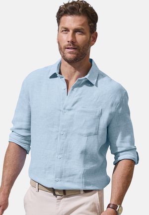 Man wearing light blue long-sleeve button-up shirt with rolled sleeves, beige braided belt, light pants, and wristwatch against a white background.