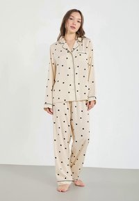 Pajama set u krem boji s uzorkom crnih srca, koji se sastoji od košulje s dugmićima i džepom te odgovarajućih hlača, izrađenih od mekog materijala.
