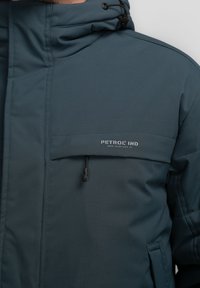 Mørk teal hættetrøje med polstret tekstur, lynlåslomme foran og snoretræk ved kraven. Logo på brystet.