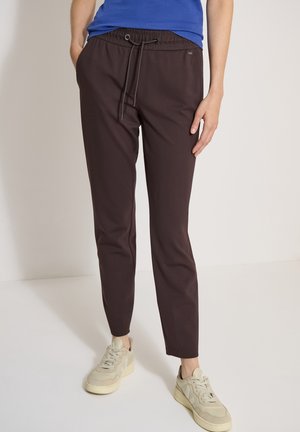 Personne portant un pantalon à cordon marron foncé, un haut bleu et des baskets beige, debout devant un fond uni.