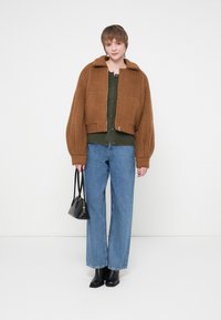 Brun cropped jakke med en tekstureret finish, grøn knapbluse og vide blå jeans. Modellen holder en lille sort håndtaske. Sorte støvler fuldender looket.