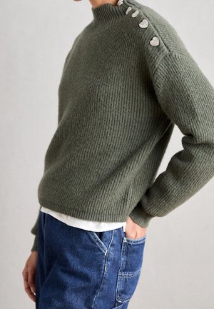 Oliivinvihreä ribbineulottu pusero korkealla kauluksella ja viisi hopeanväristä sydämenmuotoista nappia olkapäällä, yhdistetty sinisiin farkkuihin.