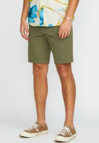 Shorts olive en coupe régulière, fabriqués dans un tissu lisse. Associés à des baskets marron avec des lacets blancs et des semelles en caoutchouc.