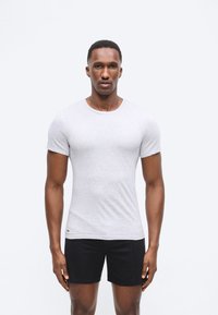 Άνδρας που στέκεται και κοιτάει μπροστά, φοράει απλό ανοιχτό γκρι τ-shirt και μαύρα σορτς μπροστά σε λευκό φόντο.