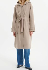 Světlé béžový dlouhý kabát s kapucí, zpřísněný v pase a s velkými kapsami. Má pásek na zavazování a druky po celé přední části.