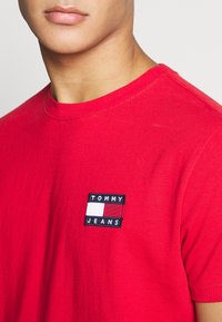 Czerwony bawełnowy t-shirt z okrągłym dekoltem, z czarną prostokątną łatą z napisem "TOMMY JEANS" oraz poziomym wzorem z kolorowych pasów.