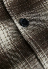 Tessuto a quadri marrone e beige con una consistenza morbida; presenta un bottone in plastica nera al centro, evidenziando la cucitura dettagliata.