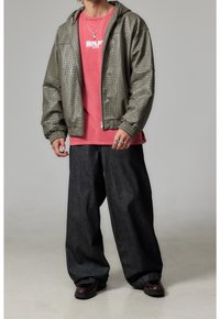 Zielona bomberka z teksturowanym wzorem i kapturem, założona na różową koszulkę, w zestawieniu z luźnymi, ciemnymi dżinsami i brązowymi butami. Zawiera widoczne elementy metalowe.