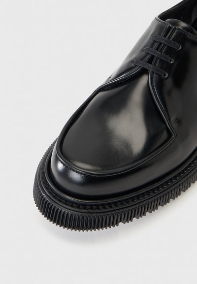 Chaussure habillée en cuir noir poli avec semelle épaisse rainurée et lacets noirs sur fond clair.
