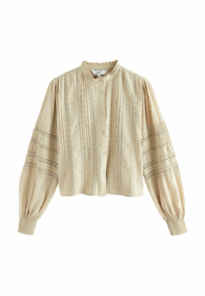 Blouse beige à boutons avec un col haut, tissu texturé, détails en dentelle et manches évasées. Présente des poignets élastiques et un design court.