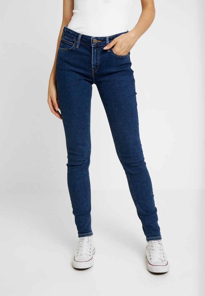 Blå denimtajta jeans med midjemedelhöjd, fem fickor och en diskret tvätt, bärs med vita sneakers.
