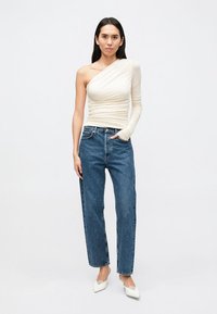 Top bege claro de um ombro só com textura franzida combinado com jeans azul de cintura alta e corte descontraído. Modelo usa saltos brancos de bico fino.