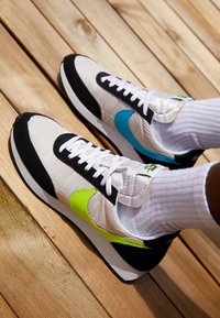 Baskets avec une tige blanche, des accents noirs et des détails en vert néon et bleu. Présente une surface texturée et un design classique à lacets.