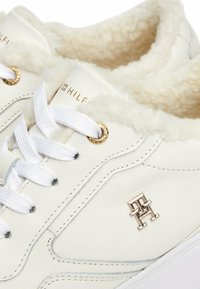 Sneaker bianche in pelle con morbido rivestimento beige. Presentano occhielli dorati, lacci bianchi e un accento metallico del logo sul lato. Design pulito e moderno.