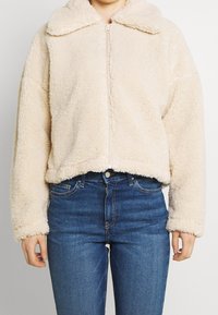 Corta giacca in ecopelliccia color crema con zip frontale, ampio colletto e vestibilità rilassata. Presenta una texture morbida e orli e polsini a costine.