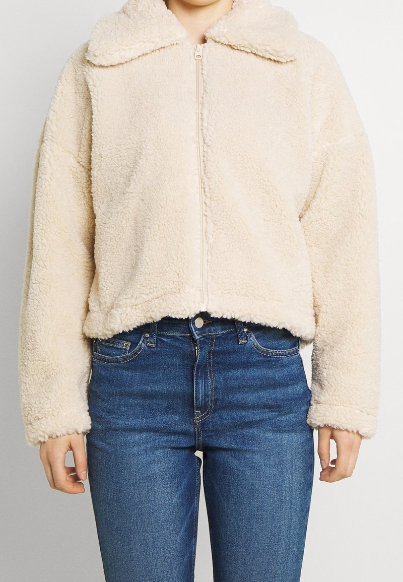 Corta giacca in ecopelliccia color crema con zip frontale, ampio colletto e vestibilità rilassata. Presenta una texture morbida e orli e polsini a costine.