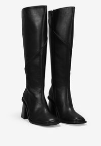 Botas de cuero negro hasta la rodilla con tacón cuadrado, que presentan un diseño de costura diagonal y cierre de cremallera en el lado interior. Textura suave.