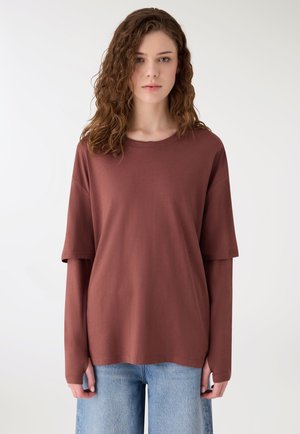 Młoda kobieta o falowanych włosach nosi warstwowe brązowe koszulki z długim i krótkim rękawem w połączeniu z jasnoniebieskimi dżinsami, stojąc na jednolitym tle.