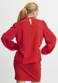 Crvena bluzica s dugim napuhanim rukavima i detaljem u obliku rupice na leđima. Izrađena od glatkog, laganog materijala. Uparena s pripijenom suknjom u istoj boji.