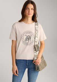Light beige t-shirt with a round graphic logo in gray, paired with blue jeans and a small gray crossbody bag featuring a patterned strap.