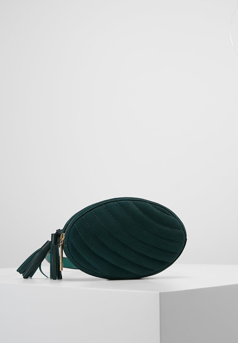 Sac bandoulière matelassé ovale vert foncé avec tirages à fermeture éclair à pompons sur une surface blanche sur fond gris.