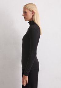 Blonde Frau in einem schwarzen Langarm-Turtleneck und einem Rock steht im Profil vor einer schlichten weißen Wand.
