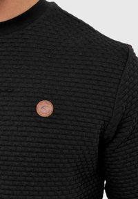 Zwarte gebreide sweatshirt met een geribbelde halslijn, voorzien van een klein bruin leren label met reliëfbranding aan de voorkant.