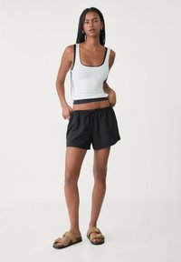 Witte aansluitende tanktop met zwarte afwerking, gecombineerd met losse zwarte shorts met een elastische tailleband. Model draagt beige sandalen.