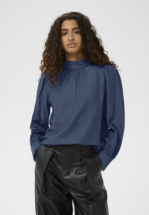 Donna con lunghi capelli ricci che indossa una blusa blu a collo alto con maniche a sbuffo e pantaloni in pelle neri, mani nelle tasche, sfondo neutro.